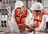 Two workers in hard hats and safety vests looking over a laptop Two workers in hard hats and safety vests looking over a laptop