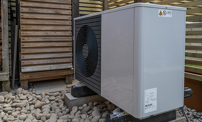 Side view of a heat pump outside in a rocky garden, near wooden fence on a sunny day.