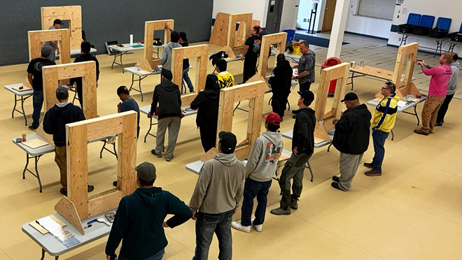 Several desks with wood frames in a large space with trainees at each desk watching an instructor show how to do a window installation as part of a high-performance building workshop