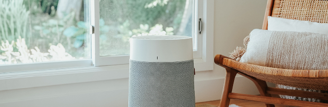 A portable air purifier in a room with the lower portion of a large window in the background on the left and on the right part of a brown wicker chair with white cushions.