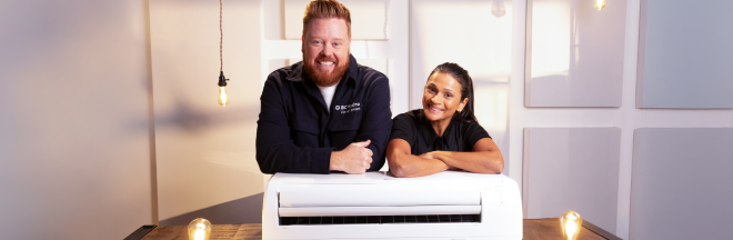Dave and Jaclyn in a room leaning cross-armed on a heat pump placed on a table. In the background are white walls and a few clear light bulbs turned on.
