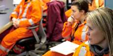 A group of workers in orange high-visibility coveralls sit in a room during a meeting or training session. A group of workers in orange high-visibility coveralls sit in a room during a meeting or training session.