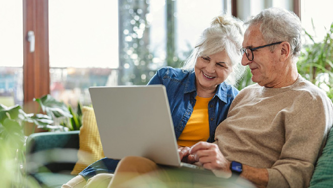 Smiling older couple sitting on a sofa looking at a laptop together in a bright living room. Smiling older couple sitting on a sofa looking at a laptop together in a bright living room.