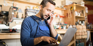 Small business owner in blue overalls uses a laptop in a tool-filled workshop.