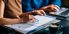 Two colleagues reviewing financial data and charts. The focus is on teamwork and analytical skills in a modern office setting with a laptop and coffee.
