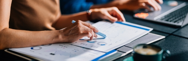Two colleagues reviewing financial data and charts. The focus is on teamwork and analytical skills in a modern office setting with a laptop and coffee.