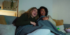 Two smiling women watch TV at home in the evening with dim lighting.