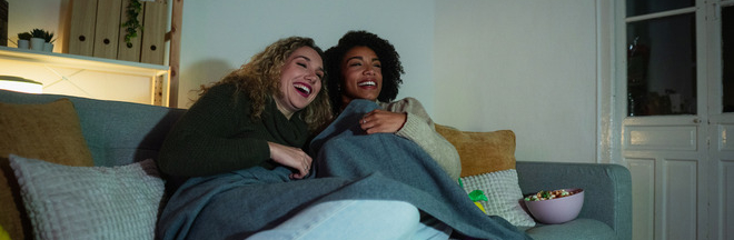 Two smiling women watch TV at home in the evening with dim lighting.