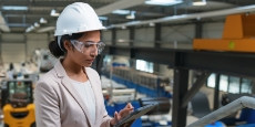 Woman at an industrial facility wearing a white safety helmet, safety glasses while working on a tablet device.
