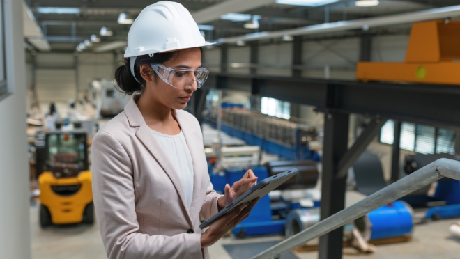 Woman at an industrial facility wearing a white safety helmet, safety glasses while working on a tablet device.