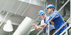 Workers discussing inside industrial facility with visible ducts and pipes.
