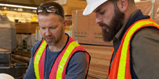Two people in safety vests are seen in a warehouse setting with stacks of wooden planks behind them.