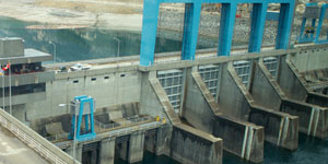 The four spillway gates of Keenlyside Dam stretches across the photo with the lifts for each gate extending upward. The reservoir is seen behind the dam. 