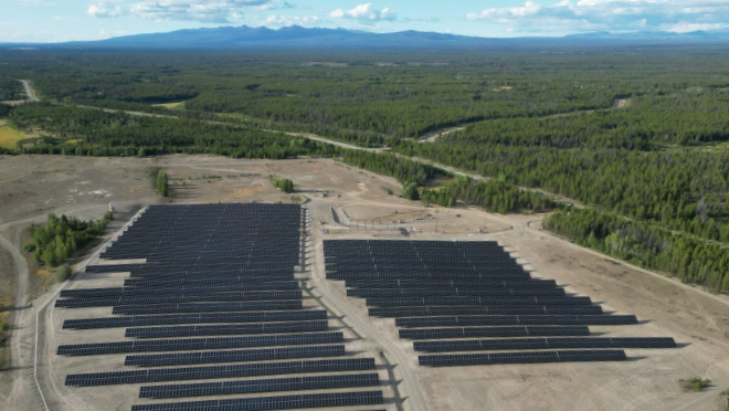 Aerial view of a solar farm with rows of solar panels on cleared land, surrounded by dense green forest.