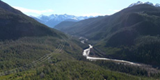 Aerial view of a winding Copper River flowing through a densely forested mountain valley and power lines crossing the landscape under a clear blue sky. Aerial view of a winding Copper River flowing through a densely forested mountain valley and power lines crossing the landscape under a clear blue sky.