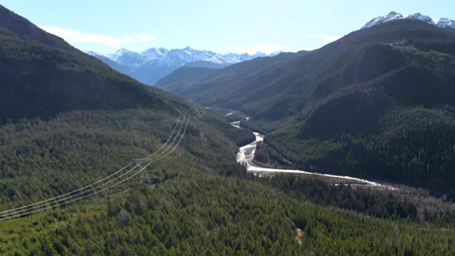 Aerial view of a winding Copper River flowing through a densely forested mountain valley and power lines crossing the landscape under a clear blue sky.
