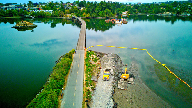 Aerial view of shoreline construction beside a causeway, with an excavator, dump truck, and containment boom in a bay.