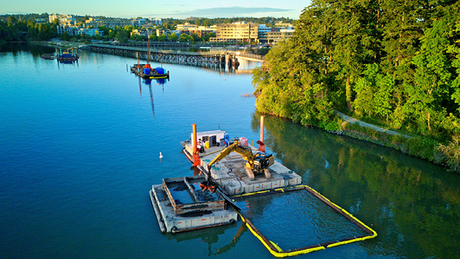 Excavator on a barge dredging in a river beside a bridge and a tree-lined shoreline within the Gorge Waterway Park, Victoria.
