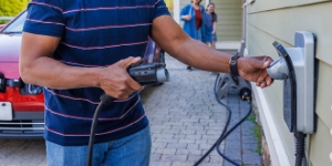 A man plugs an electric charger. The background shows his interracial family walking around and approaching.