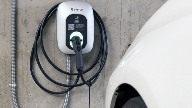 A wall-mounted SWTCH electric vehicle charging station with coiled charging cable in an indoor parking garage, next to a white EV.