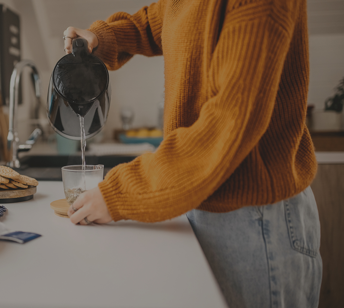Person in dark orange sweater and faded jeans standing at a counter pouring water from a kettle into a cup.