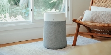 A portable air purifier in a room with the lower portion of a large window in the background on the left and on the right part of a brown wicker chair with white cushions.