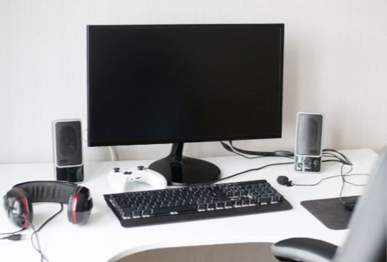 A wide-screen computer monitor on a white desk with two speakers, a pair of over-the-ear headphones, a game controller and a keyboard.