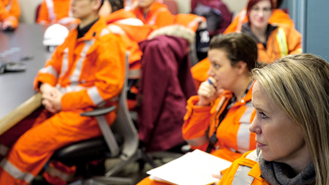 A group of workers in orange high-visibility coveralls sit in a room during a meeting or training session.