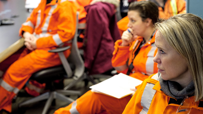 A group of workers in orange high-visibility coveralls sit in a room during a meeting or training session.