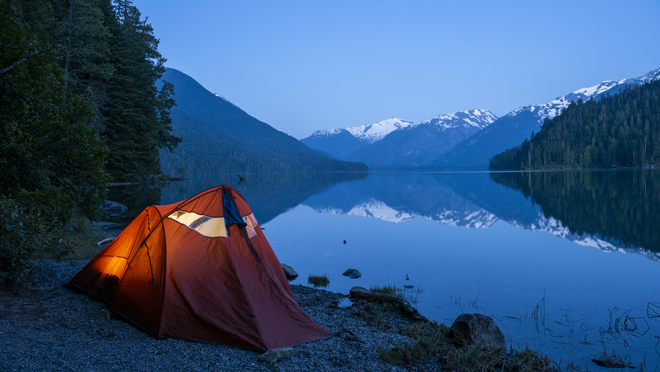 Illuminated camping tent beside a calm mountain lake with snow-capped peaks reflected in the water.